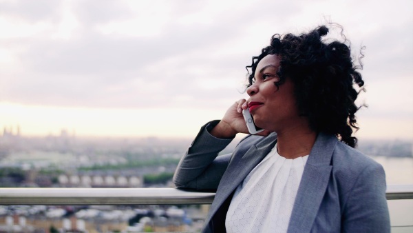 A portrait of black businesswoman standing on a terrace against London rooftop view panorama, making a phone call. Copy space.