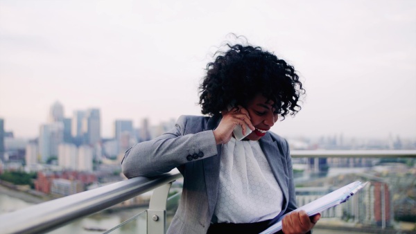 A portrait of black businesswoman standing on a terrace against London rooftop view panorama, making a phone call. Copy space.