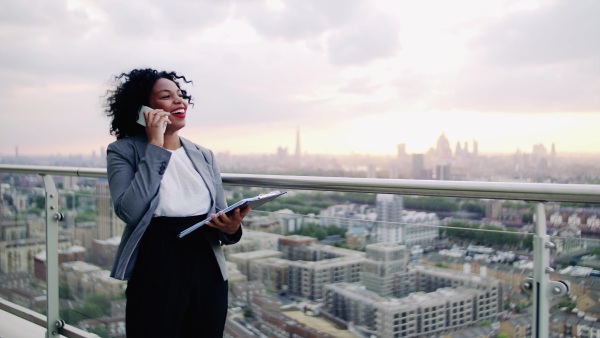 A portrait of black businesswoman standing on a terrace against London rooftop view panorama, making a phone call. Copy space.