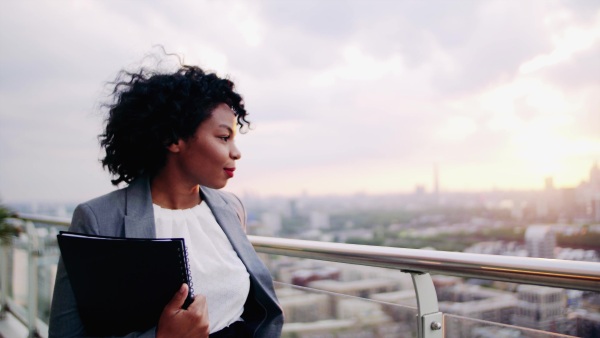 A portrait of black businesswoman walking on a terrace at sunset, holding notes. London rooftop view panorama in the background. Copy space.