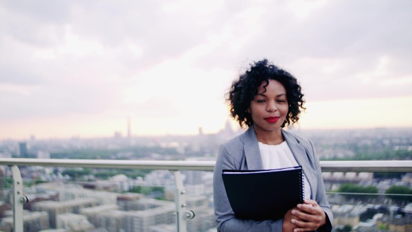 A portrait of black businesswoman standing on a terrace against London rooftop view panorama, holding notes. Copy space.