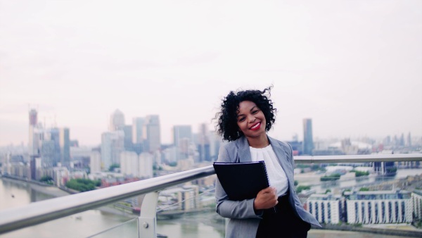 A portrait of black businesswoman standing on a terrace against London rooftop view panorama, holding notes. Copy space.