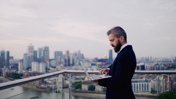 A portrait of businessman holding laptop standing against London rooftop view panorama, typing. Copy space.