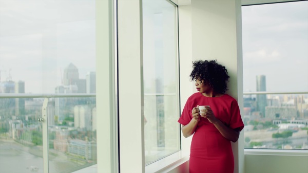 A portrait of woman standing by the window against London rooftop view panorama, holding a cup of coffee. Copy space.