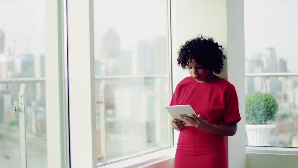 A portrait of woman standing by the window against London rooftop view panorama, using tablet. Copy space.