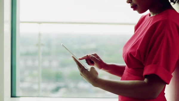 An unrecognizable woman standing in an office by the window in an office, using tablet. Copy space. Slow motion.