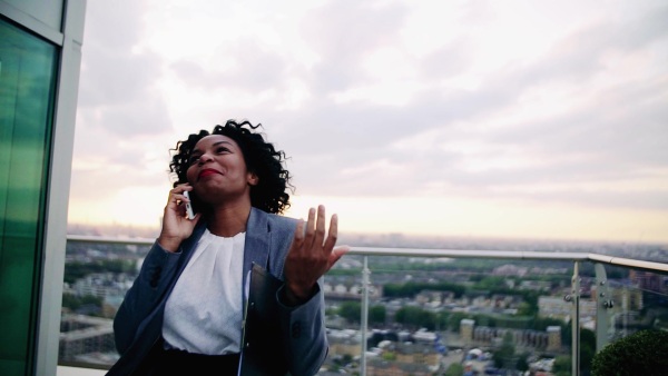 A portrait of black businesswoman standing on a terrace against London rooftop view panorama, making a phone call. Copy space. Slow motion.