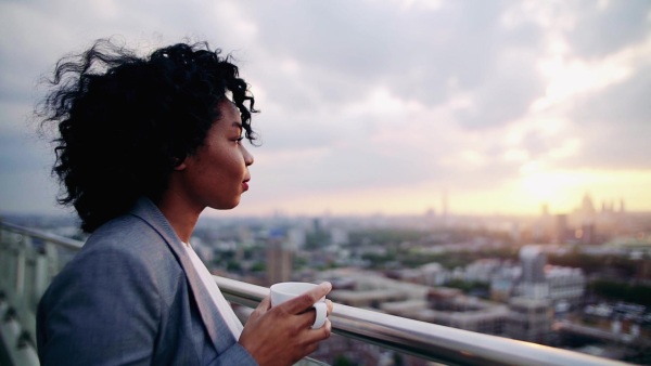 A portrait of black businesswoman standing on a terrace against London rooftop view panorama, drinking coffee. Copy space. Slow motion.