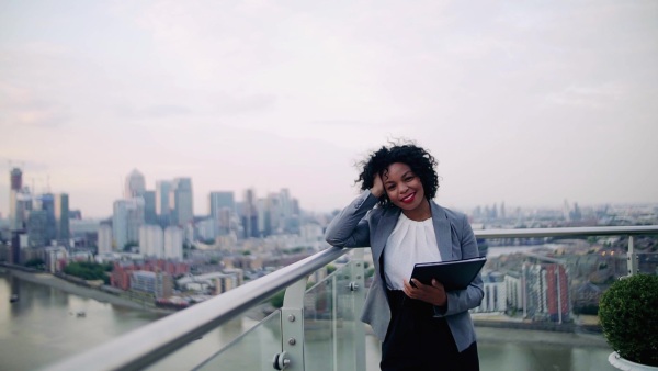 A portrait of black businesswoman standing on a terrace against London rooftop view panorama, holding notes. Copy space. Slow motion.