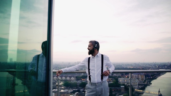 A cheerful businessman with headphones standing against London view panorama, listening to music. Slow motion.