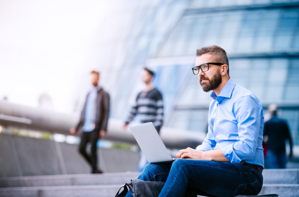 Hipster manager sitting on stairs on sunny day, working on laptop, London, City Hall