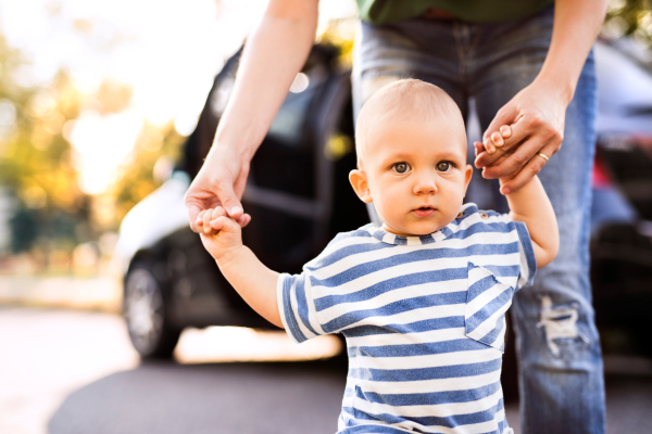 Unrecognizable young mother helping her little baby boy walk. First steps, near a car.