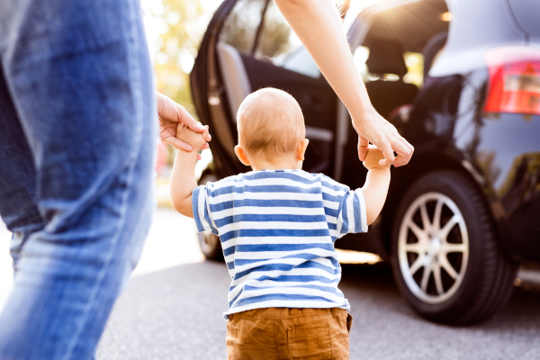 Unrecognizable young mother helping her little baby boy walk. First steps, near a car. Rear view.