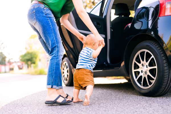 Unrecognizable young mother helping her little baby boy walk. First steps, near a car.