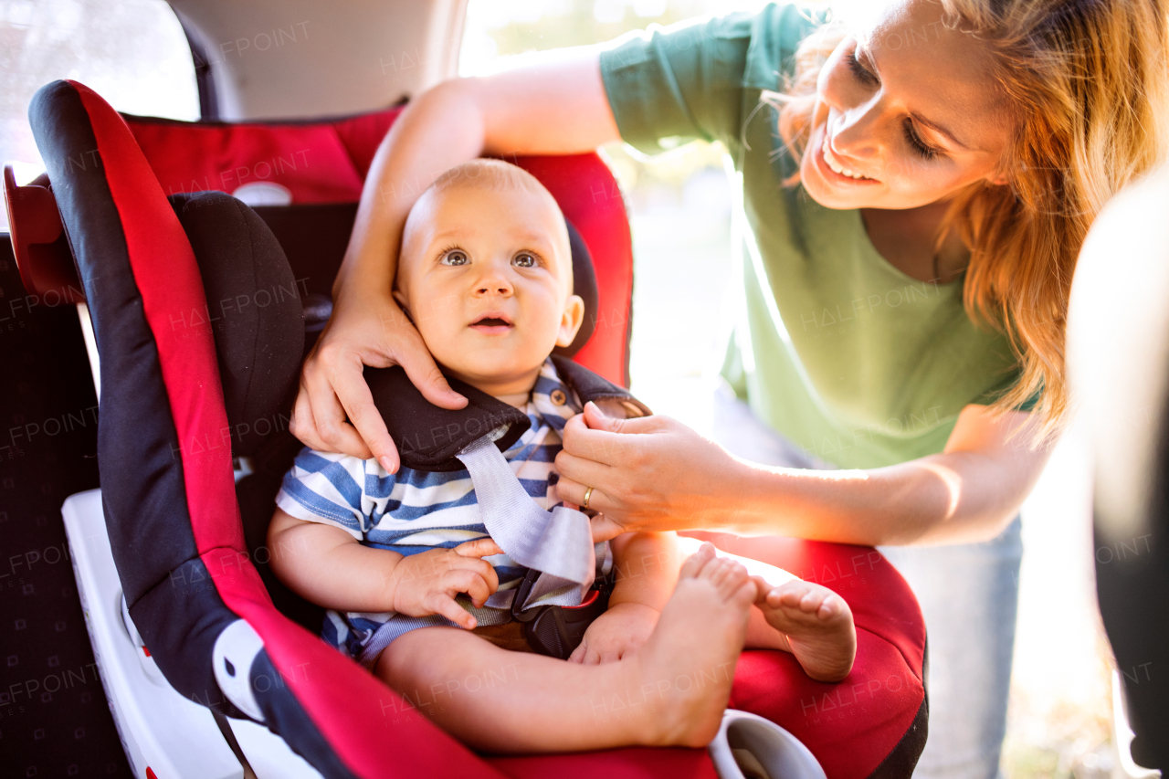 Young mother putting her little baby boy in the car seat, fastening seat belts.