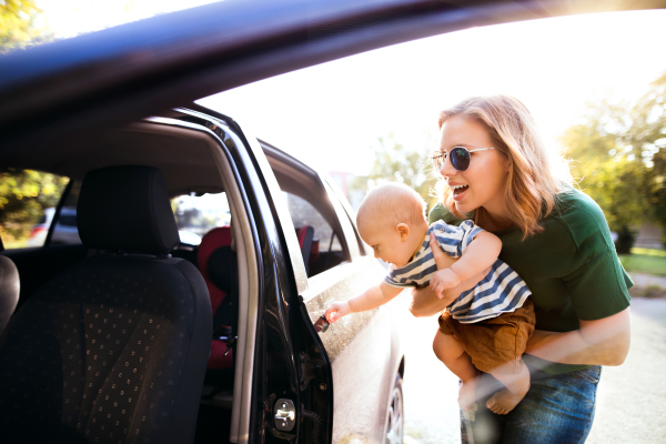 Young mother holding her little baby boy in the arms getting into the car. Shot through glass.