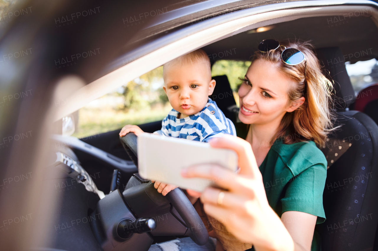 Young mother with her little son in the car. A woman and baby boy pretending to be driving. Woman taking photo with a smartphone.