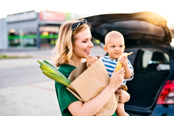 Beautiful young mother with her little baby son in front of a supermarket, holding paper shopping bag. Woman with a boy standing by the car.