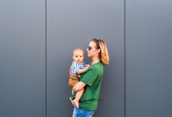 Young woman with a baby boy standing against the wall. Beautiful mother holding her son in arms.