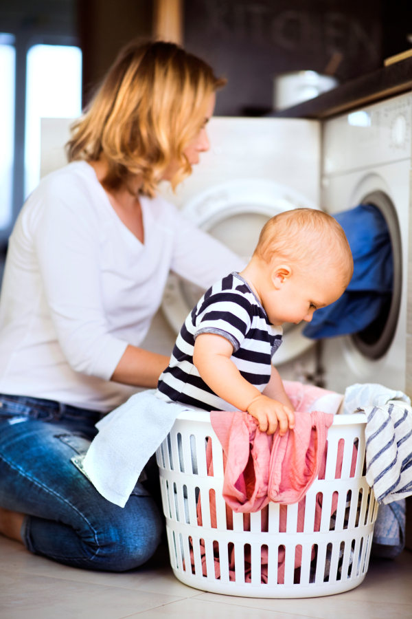 Young mother with a baby son doing housework. Beautiful woman and baby boy doing laundry. Baby boy in the laundry basket.