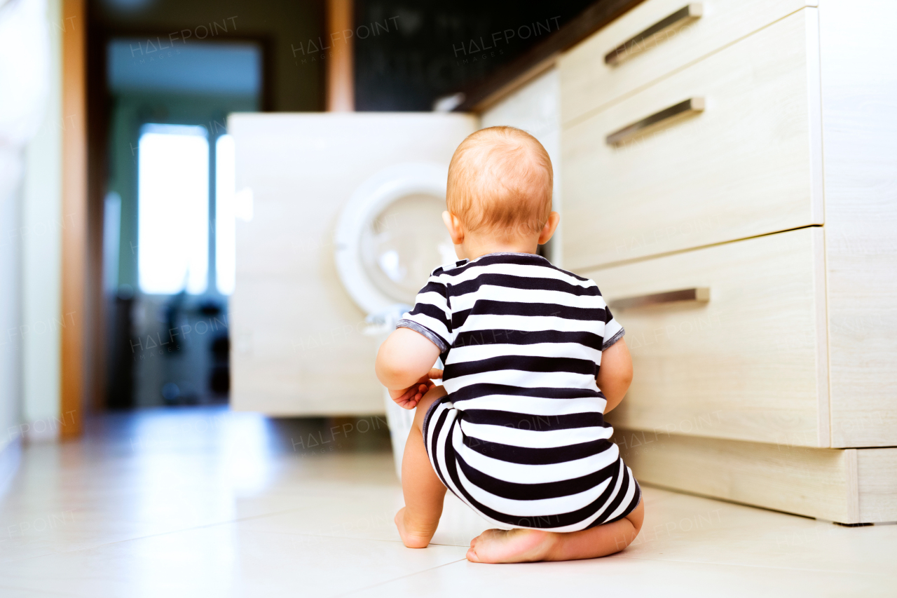 Cute baby boy by the washing mashine in the kitchen. Laundry basket on the floor. Rear view.