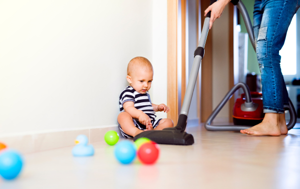 Unrecognizable young mother with a baby son doing housework. Woman hoovering and baby boy sitting on the floor.