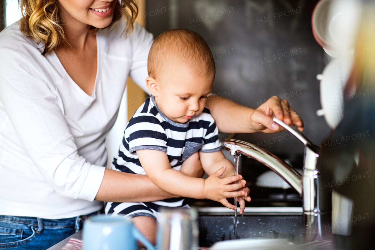 Unrecogniozable young mother with a baby son doing housework. Beautiful woman and baby boy washing up the dishes.