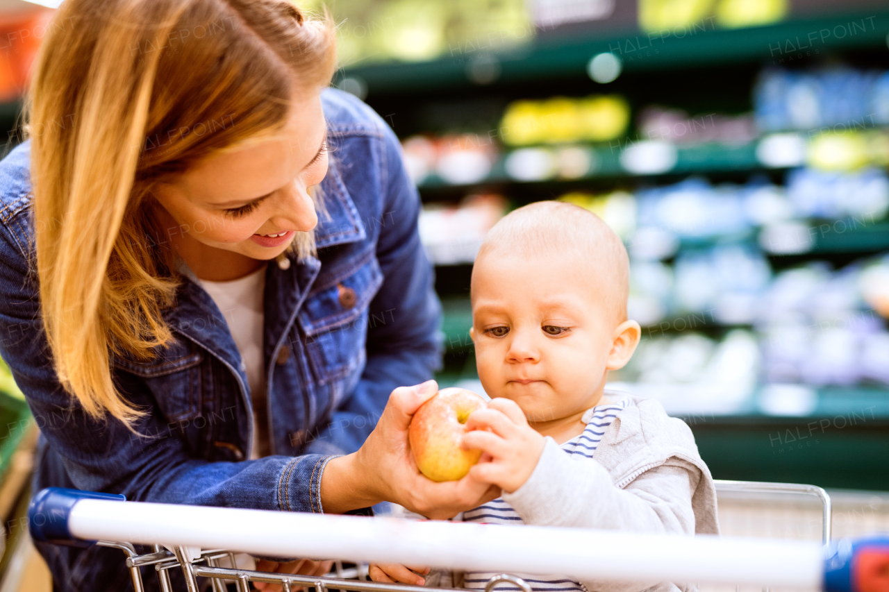 Beautiful young mother with her little baby boy at the supermarket, shopping. Woman giving her son an apple. Close up.