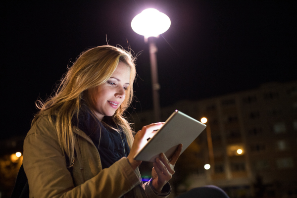 Beautiful woman in the city at night holding tablet, reading something.