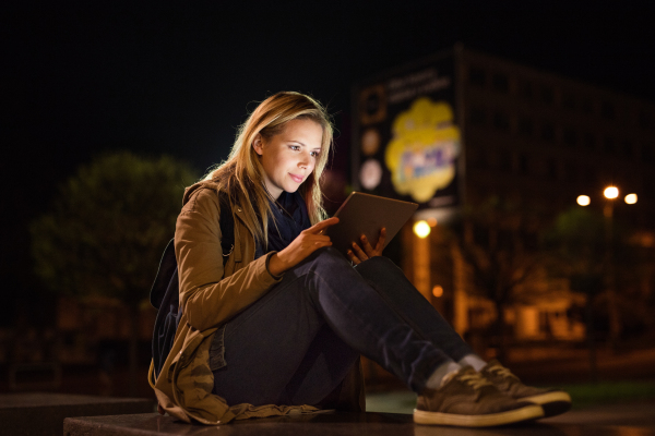 Beautiful woman in the city at night sitting on the ground, holding tablet, reading something.