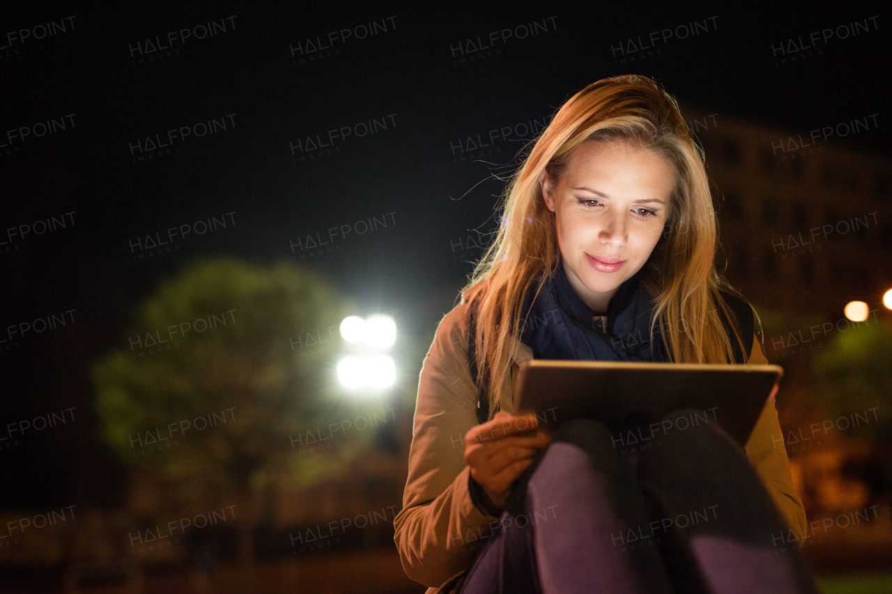 Beautiful woman in the city at night holding tablet, reading something.