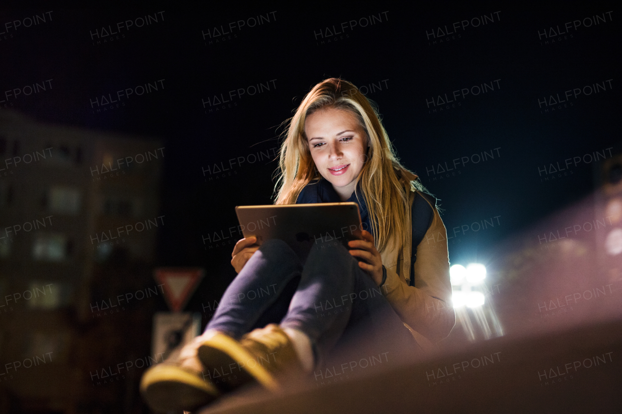 Beautiful woman in the city at night sitting on the ground, holding tablet, reading something.