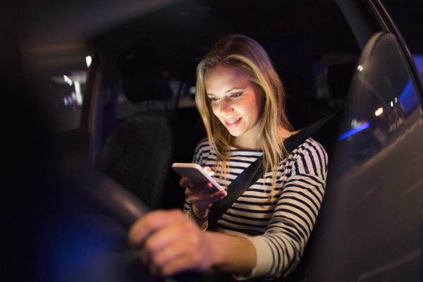 Beautiful young woman in a city in her modern car at night, holding smart phone, texting.