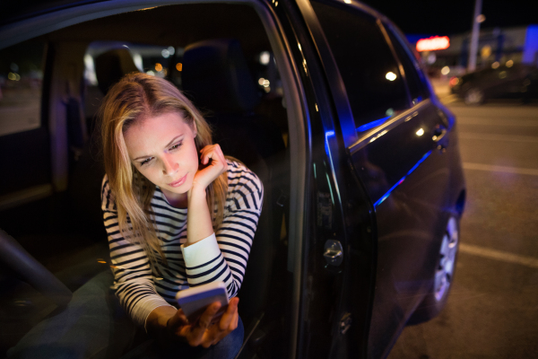 Beautiful young woman in a city in her modern car at night, holding smart phone, texting.