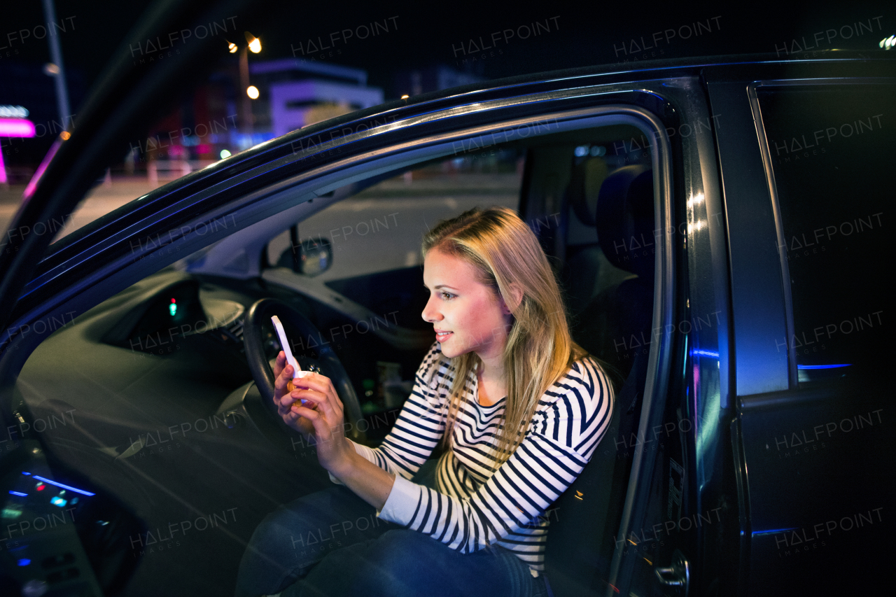 Beautiful young woman in a city in her modern car at night, holding smart phone, texting.