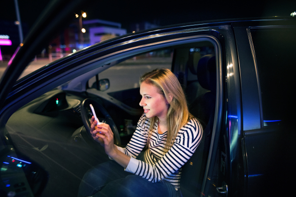 Beautiful young woman in a city in her modern car at night, holding smart phone, texting.