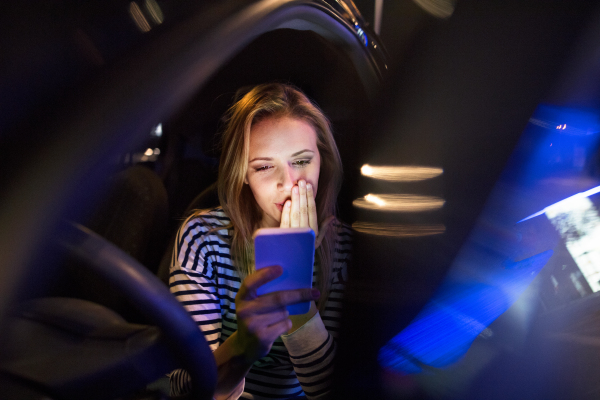 Beautiful young woman in a city in her modern car at night, holding smart phone, reading text message.