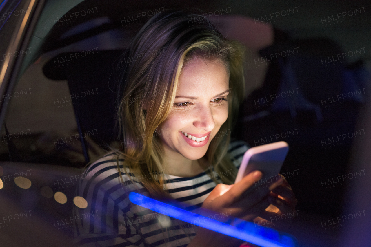 Beautiful young woman in a city in her modern car at night, holding smart phone, texting.