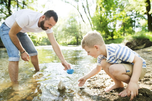 Young father with little boy playing with paper boats at river bank, sunny spring day.