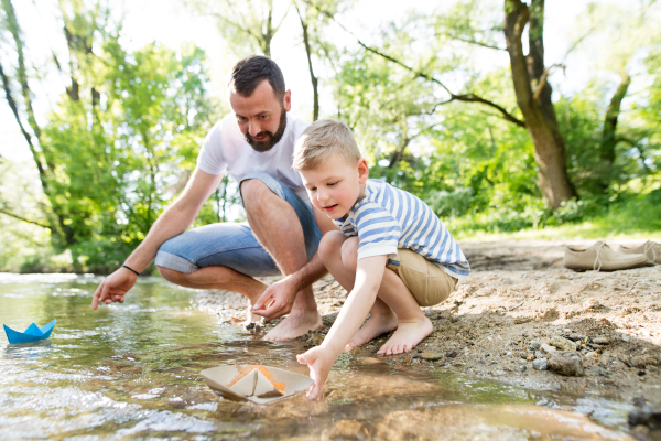 Young father with little boy playing with paper boats at river bank, sunny spring day.