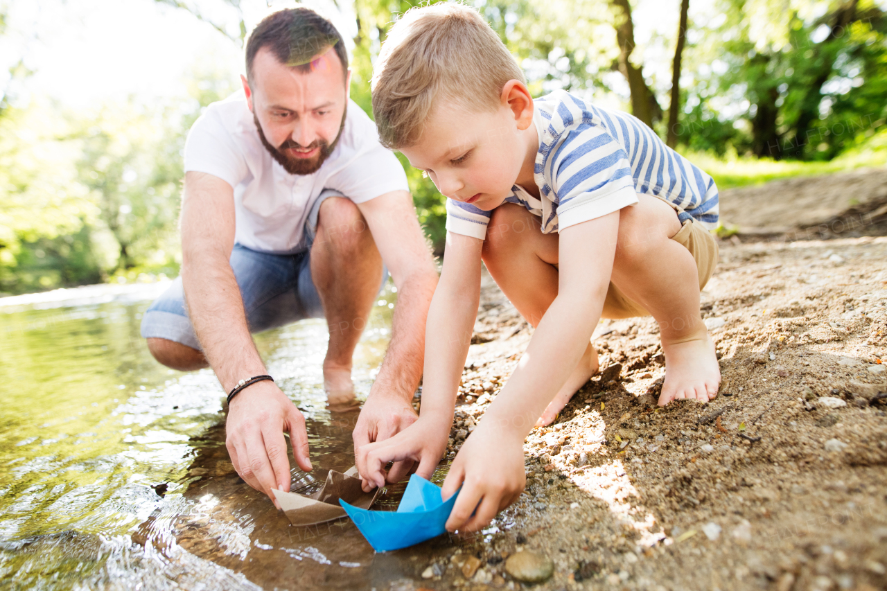 Young father with little boy playing with paper boats at river bank, sunny spring day.