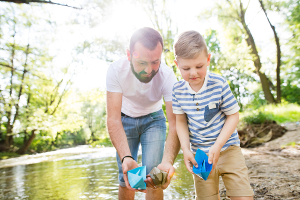 Young father with little boy playing with paper boats at river bank, sunny spring day.