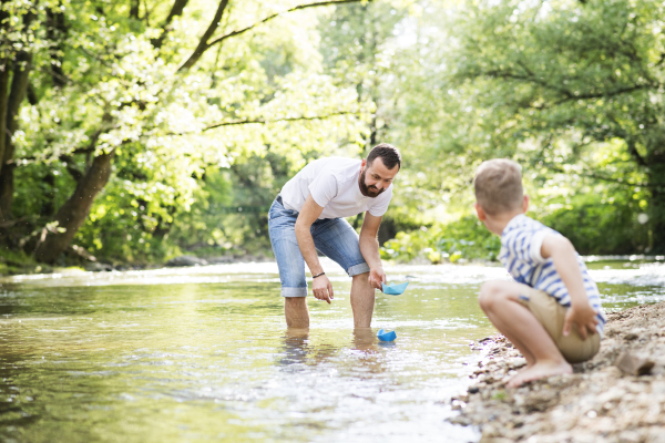 Young father with little boy playing with paper boats at river bank, sunny spring day.