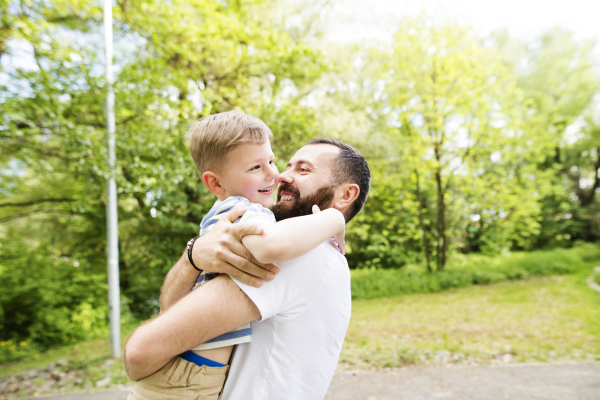 Young father with his little son in green woods holding him in the arms, hugging him, sunny day.