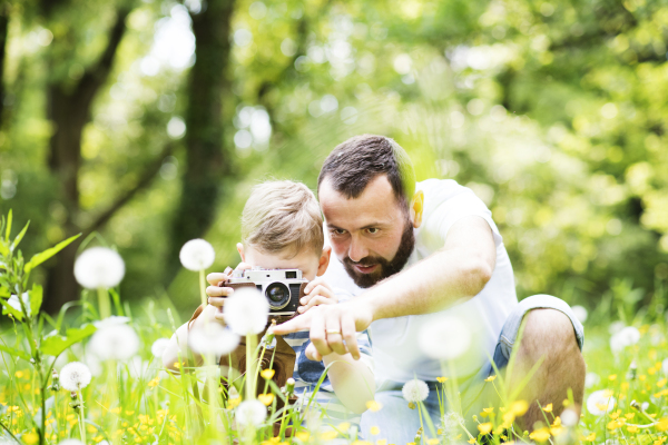 Young hipster father outdoors with his little son with camera, taking pictures of nature. Sunny summer day.