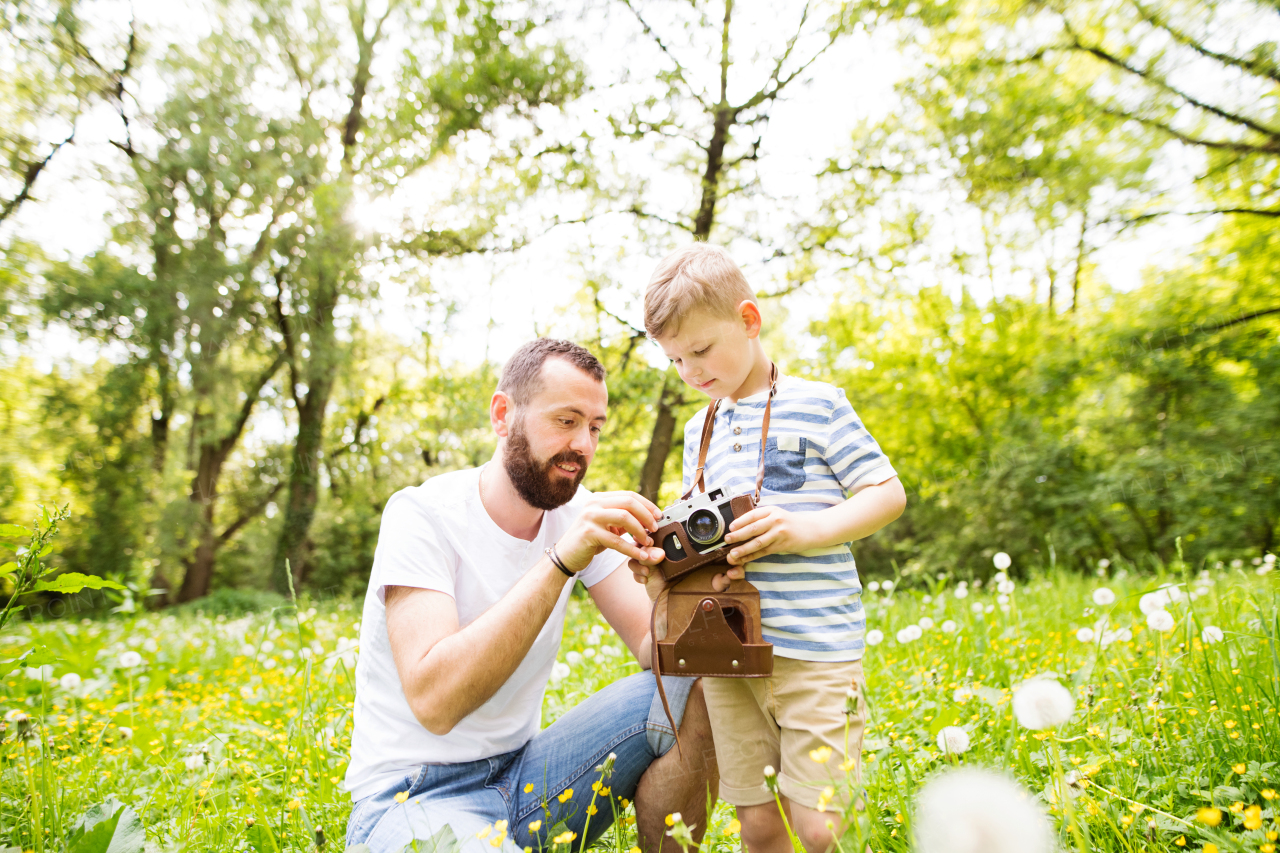 Young hipster father outdoors with his little son with camera, explains and shows him how to use it. Sunny summer nature.