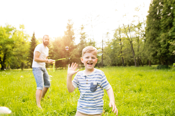 Young hipster father with little boy outside blowing bubbles, having fun, sunny summer day.