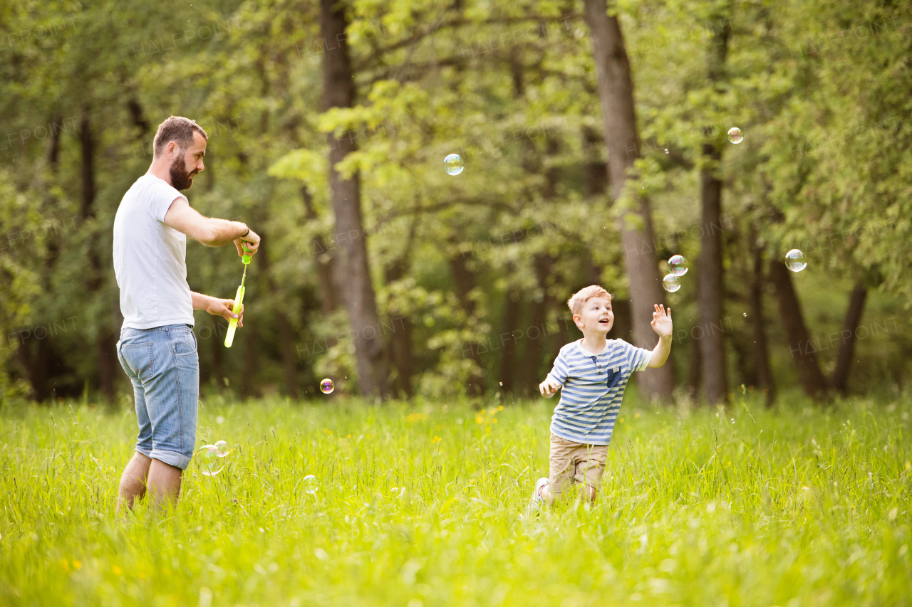 Young hipster father with little boy outside blowing bubbles, having fun, sunny summer day.