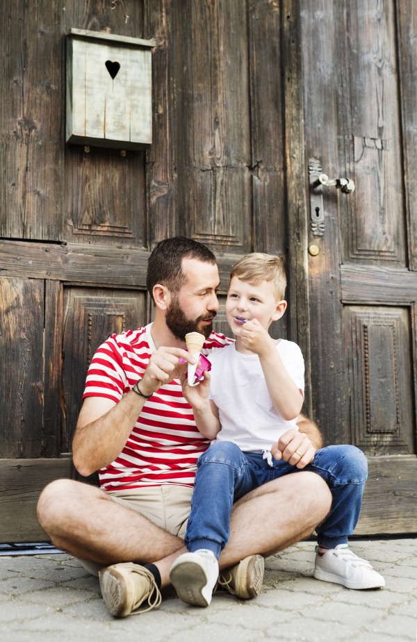 Young father and his little son eating ice cream, sitting on the floor in front of old big door. Sunny summer day.