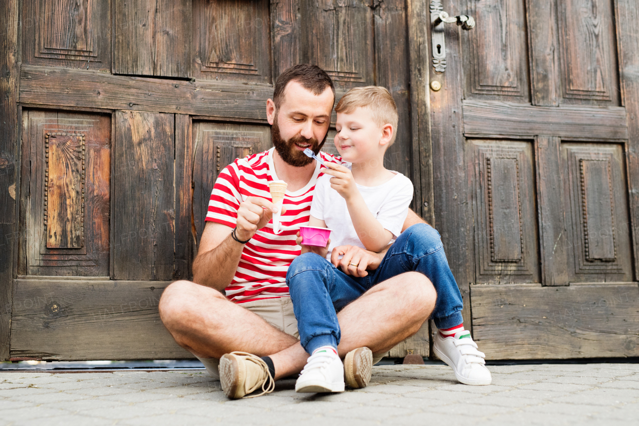 Young father and his little son eating ice cream, sitting on the floor in front of old big door. Sunny summer day.